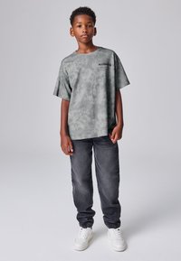 Chlapec stojaci s uvoľnenými rukami, oblečený v zelenom batikovanom tričku s logom "ALLSAINTS", tmavých džínsoch a bielych teniskách na jednoduchom pozadí.