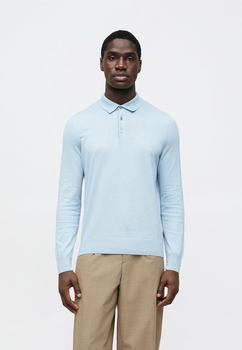 Gaiši zils polo krekls ar garām piedurknēm un apkakli, ar teksturētu apdari un trīs pogu aizdari. Valkāts ar bēšām biksēm.