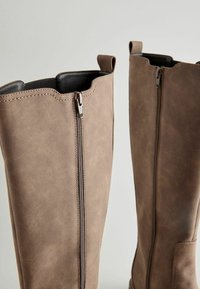 Brown leather knee-high boots with a smooth texture, featuring a front zipper and pull tabs at the tops for easy wearing.