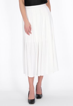 Žena v bílé plisované midi sukni, černém tílku bez rukávů a černých vysokých podpatcích stojící na bílém pozadí.