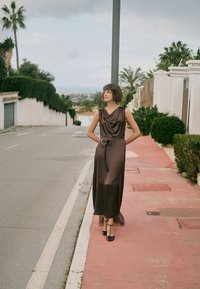 Femme en longue robe sombre et talons, debout sur un trottoir près d'un lampadaire dans une rue calme avec de la verdure et des palmiers.