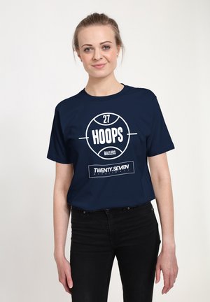 Jonge vrouw met lichtbruin haar in een knot, gekleed in een marineblauw T-shirt met de tekst "27 Hoops Ballers" en een zwarte broek, staande tegen een witte achtergrond.