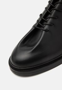 Black leather shoe with a rounded toe, smooth texture, and black laces. Features a slightly raised sole with a minimalistic design.