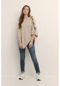 Smėlio spalvos pončas su raukiniais, stačiakampio formos, minkštos tekstūros, derinamas su mėlynais džinsais ir baltais sportbačiais, stovintis prieš neutralią foną.