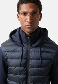 Marineblå puffervest med lynlåslukning, der har vandrette quiltede sektioner, kombineret med en hættetrøje under. Briller med klare stel.