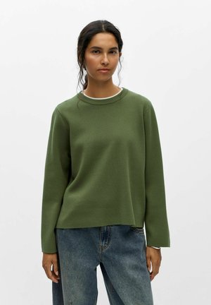 Kvinde står iført en simpel olivengrøn langærmet sweater og blå jeans mod en ensfarvet hvid baggrund.