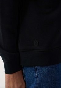 Sudadera negra, hecha de una tela suave, con puños y dobladillo acanalados. Muestra un pequeño parche de logo negro en el lado inferior.