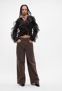 Blusa nera trasparente con maniche a volant, abbinata a pantaloni larghi marroni. L'outfit presenta un design corto e un nastro al collo.