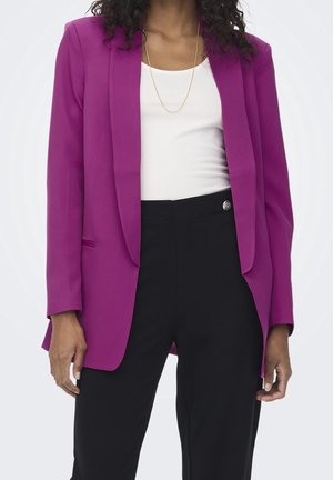 Frau trägt einen hellvioletten Blazer, weißes Oberteil, schwarze hoch taillierte Hose und eine dünne goldene Halskette vor hellem Hintergrund.