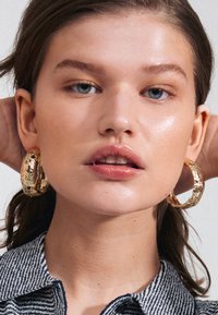 Jeune femme avec des yeux bleus et un maquillage léger portant des boucles d'oreilles créoles texturées en or et une chemise à col rayée, les mains derrière la tête.
