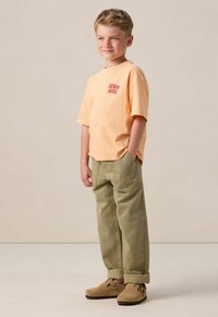 Lichtoranje T-shirt met korte mouwen met "VENICE BEACH" in het rood erop gedrukt, gecombineerd met olijfgroene broek en tan instap schoenen.