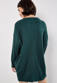 Vrouw met kort, donker haar, gekleed in een losse, gestructureerde donkergroene trui jurk met lange mouwen, die met de rug naar de camera is gekeerd tegen een effen witte achtergrond.