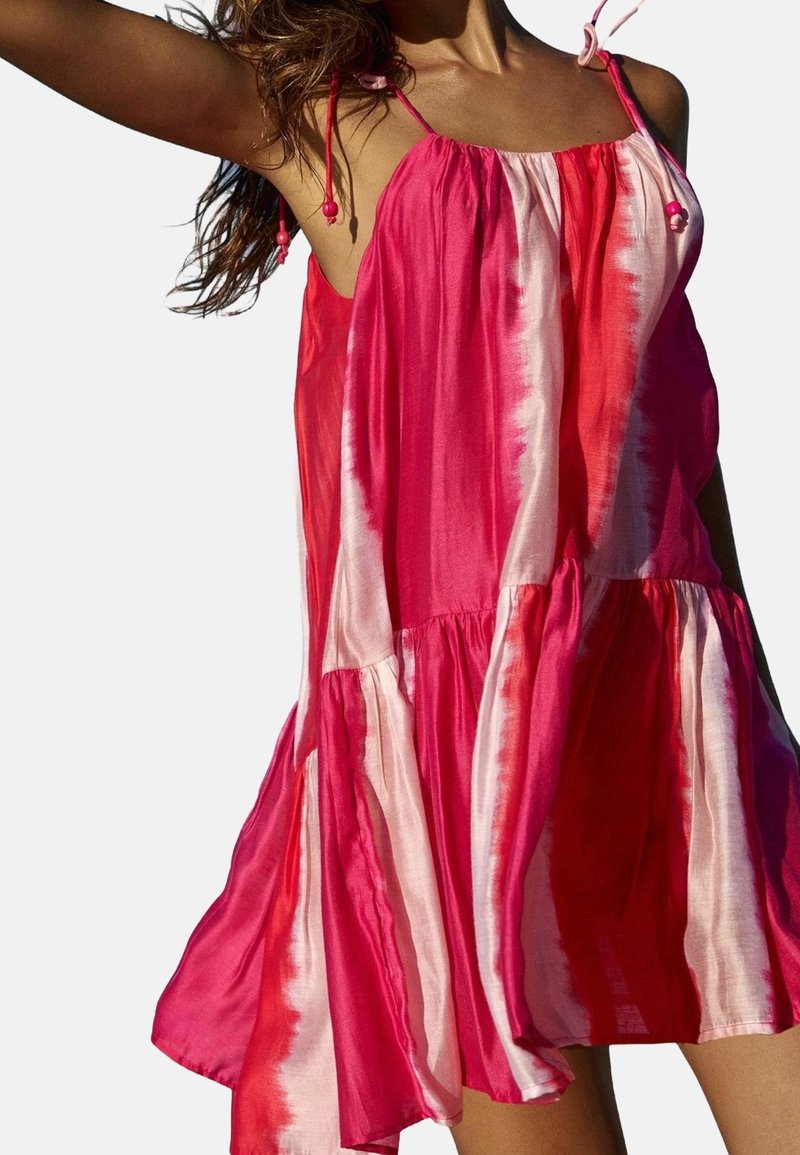Femme portant une robe ample sans manches avec des rayures verticales roses et rouges et de fines bretelles sur fond clair uni.