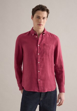 Hombre joven con una camisa roja de manga larga abotonada y vaqueros oscuros, de pie frente a un fondo liso de color gris claro.