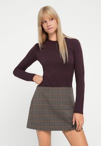 Langärmliges lila Oberteil, kombiniert mit einem karierter Rock in braunen, beigen und grünen Tönen, mit geradem Saum und seitlichen Taschen.