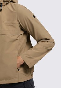 Giacca impermeabile beige con tasca frontale con zip, cappuccio regolabile e polsini elastici. Presenta un logo sul petto e dettagli con bottoni neri.