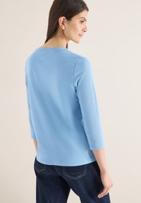 Femme de profil portant un haut bleu clair à manches trois quarts et un jean bleu foncé sur un fond beige.
