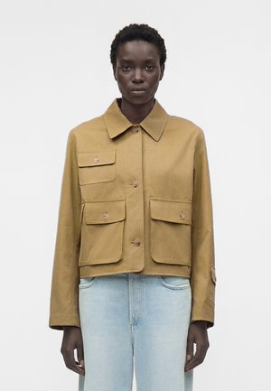 Femme aux cheveux courts et bouclés portant une veste beige boutonnée avec quatre poches à rabat à l’avant et un jean bleu clair, sur fond uni.