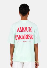 Camiseta de algodón verde menta con gran texto rojo que dice "AMOUR PARADISIO" en la parte posterior, con pequeños detalles de marca debajo. Manga corta.