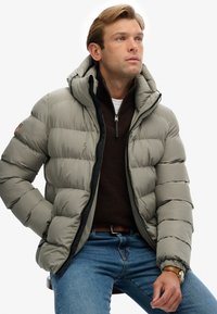 Lys grå pufferjakke med hætte, der har et quiltet design og lynlås foran. Båret over en mørkebrun zip-up trøje og blå jeans.