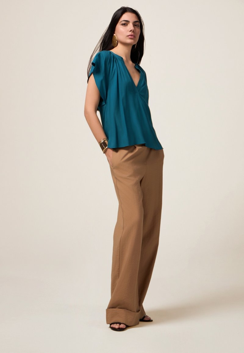 Blusa turchese con scollo a V e maniche corte abbinata a pantaloni beige larghi. Tessuto liscio con leggero lucido, dettagli minimi e vestibilità comoda.