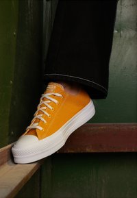 Keltainen kangaskengät valkoisella kumipohjalla ja nauhoilla, pyöreä kärki ja teksturoitu kärkitakki. Näkyvät saumat ja logo sivussa.