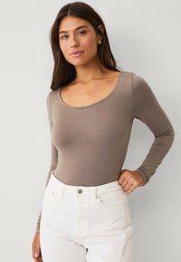 Bodysuit s dlouhým rukávem vyrobený z elastického světle hnědého materiálu s kulatým výstřihem, doplněný o bílé džíny s vysokým pasem. Jednoduchý a přiléhavý design.