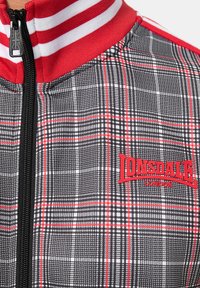 Lonsdale London jaka ar melnu un baltu šķautņu rakstu ar sarkaniem akcentiem, ar rāvējslēdzēju un sarkanu logo uz krūtīm.