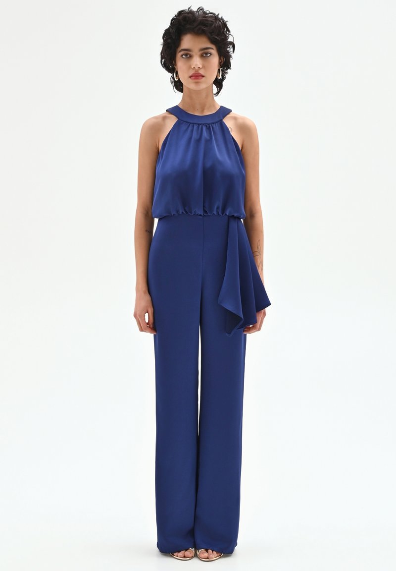 Granatowy kombinezon z odkrytymi plecami i wiązaniem na szyi, z marszczonym górnym pasem i szerokimi nogawkami. Posiada drapowany detal z jednej strony. Gładka, satynowa tekstura.