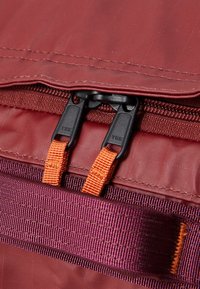 Détail d'un sac à dos en tissu rouge montrant une double fermeture éclair noire avec des tirants orange, et une sangle tissée violette avec un aspect texturé.