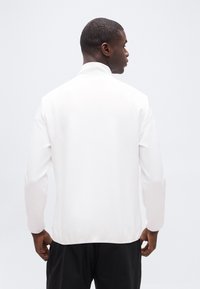 Άνδρας με κοντά μαλλιά φορώντας ένα απλό λευκό μακρυμάνικο μπλουζάκι και μαύρο παντελόνι, γυρισμένος προς τα πίσω σε ανοιχτό γκρι φόντο.