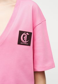 Haut rose à manches courtes avec un décolleté en V profond et un patch carré noir affichant le logo rose "Just Cavalli" sur la poitrine gauche.