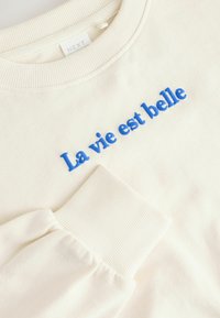 Kremowy sweatshirt z niebieskim haftowanym napisem "La vie est belle". Posiada żebrowane mankiety i dekolt. Miękka tekstura materiału.