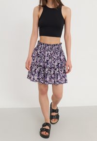 Młoda kobieta ubrana w bezrękawnikową czarną bluzkę crop, warstwową spódnicę w kwiaty w kolorze purpurowym oraz czarne sandały na klamrę na jasnym jednolitym tle.
