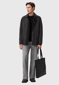 Homme en veste noire, pull noir, jean gris et chaussures noires tenant un grand sac fourre-tout noir, regardant légèrement sur le côté.