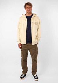 Grosse veste zippée beige avec un logo, assortie à un pantalon cargo vert olive et des baskets noires. Comprend des cordons de serrage et des poignets côtelés.