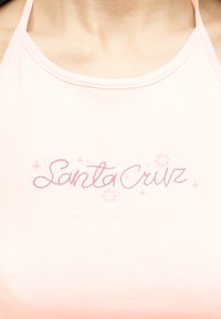 Κοντινή λήψη ενός ανοιχτορόζ τοπ με λεπτές τιράντες και την επιγραφή "Santa Cruz" σε ροζ καλλιγραφικά γράμματα με μικρές διακοσμήσεις αστέρια.