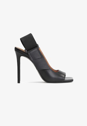 Mustat korkokengät sandaalit neliskulmaisen varpaansa kanssa, leveällä elastisella nilkkahihnalla ja nahkapinnalla, esitetty sivusta valkoista taustaa vasten.