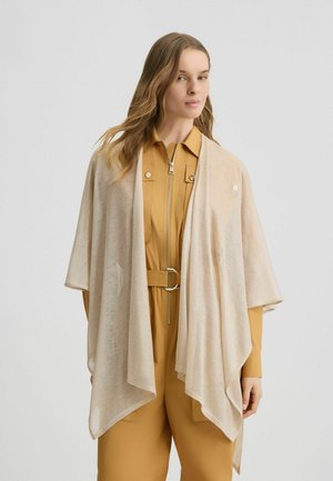 Châle beige léger au design ouvert, avec une texture douce et des manches drapées, porté sur une combinaison moutarde avec une ceinture.