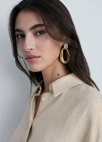 Gold hoop earring with a textured surface and rectangular crystal accent. Model wears a light beige collared shirt with a soft texture.