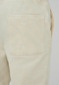 Pantalon en velours côtelé beige clair avec une texture verticale nervurée prononcée, comportant une grande poche arrière et un design minimaliste.