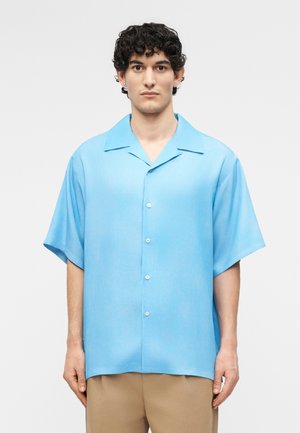 Άνδρας με σγουρά μαλλιά φοράει ανοιχτό γαλάζιο κοντομάνικο πουκάμισο με κουμπιά και μπεζ παντελόνι, στέκεται μπροστά σε απλό φόντο.