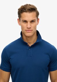 Polo shirt blu navy con maniche corte, colletto alzato, pattina con due bottoni e accento a righe sottili sotto il colletto.