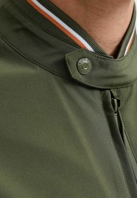 Veste vert olive avec un col haut et un accent rayé orange et blanc. Comprend une fermeture à boutons-pression et une fermeture éclair sur le côté.