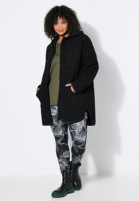 Mujer con abrigo negro con capucha, top verde oliva, leggings florales en blanco y negro y botas negras, de pie frente a un fondo blanco.