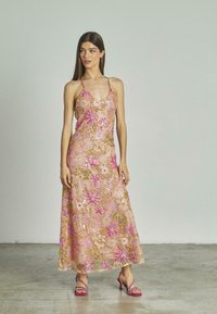 Il vestito lungo presenta un intricato ricamo floreale in rosa e oro, paillettes, spalline sottili e una silhouette fluida; abbinato a sandali con tacco rosa.