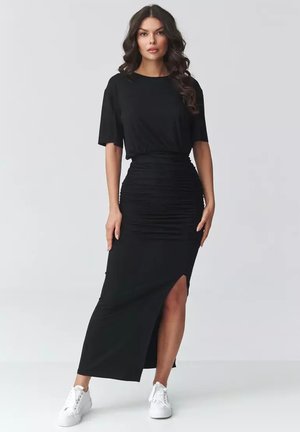 Kobieta stoi, ubrana w czarny top z krótkim rękawem i czarną spódnicę z wysokim stanem z marszczeniami oraz rozcięciem z boku, do tego białe trampki.