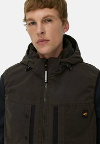Dunkelgrüne Kapuzenweste mit einem Reißverschluss, zwei vorderen Taschen mit schwarzer Einfassung und einem Logopatch. Glatte, matte Textur.