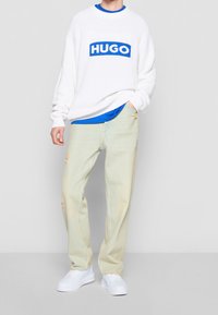 Biały sweter z dzianiny z niebieskim logo "HUGO", noszony na niebieskiej koszuli. Dopełnieniem stroju są jasnżółte, postrzępione dżinsy oraz białe sneakersy.