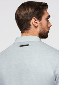 Uomo con capelli castani corti e barba, che indossa una polo grigio chiaro a trama, visto dalla parte posteriore e destra.